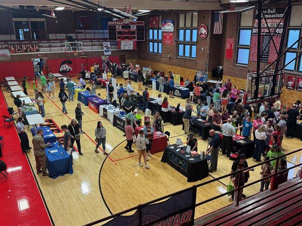Students and families wander throughout a gym. There are tables set up in four lines.